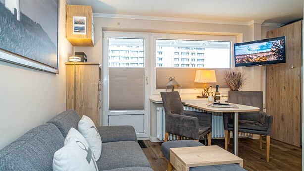 A modern living room with a grey sofa, a wooden coffee table, and a dining area with a round table and chairs. A flat-screen TV is mounted on the wall. - Strandmuschel App. 309 - Kleine, Geschmackvoll Eingerichtete Wohnung in Westerland, Direkt am (Sylt)