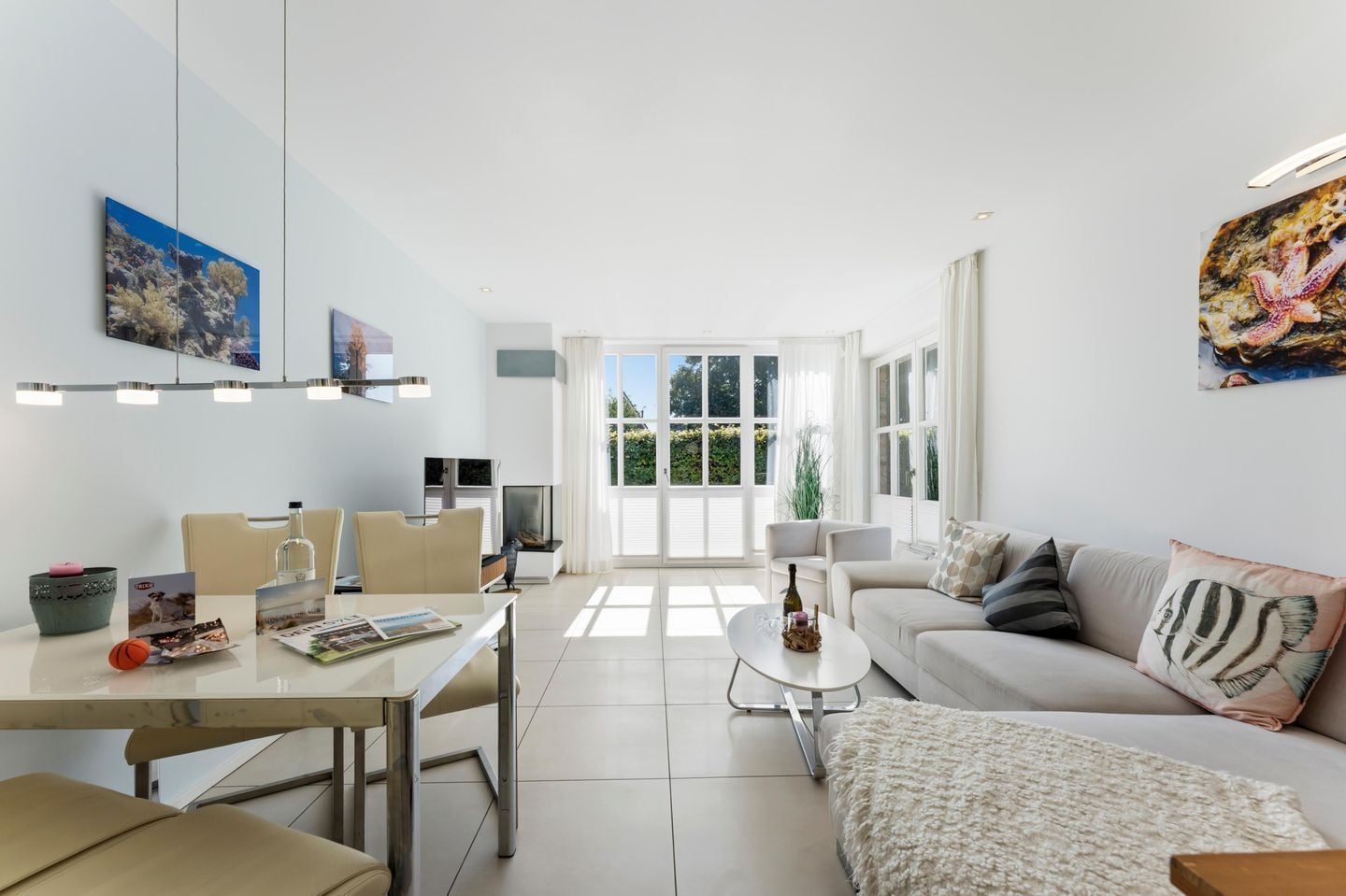 A modern living room with a sofa, coffee table, and dining area. There is a large window providing natural light and a view of greenery outside.