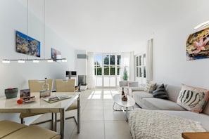 A modern living room with a sofa, coffee table, and dining area. There is a large window providing natural light and a view of greenery outside.