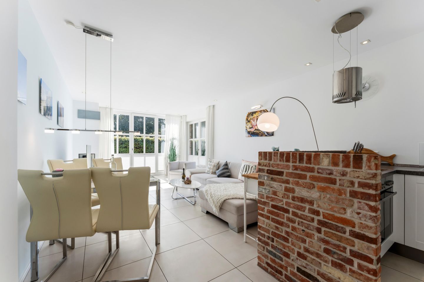 A modern kitchen with a brick accent wall, a dining area with chairs and a table, and a living area with a sofa and a lamp.