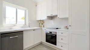 A modern kitchen with white cabinets, a dishwasher, an oven, and a window providing natural light. - Little Manly 3 bedroom Oasis (Manly)