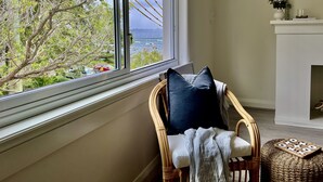 A room with a view of the sea through a large window, a wicker chair with a cushion, and a small ottoman with a wooden puzzle. - Little Manly 3 bedroom Oasis (Manly)
