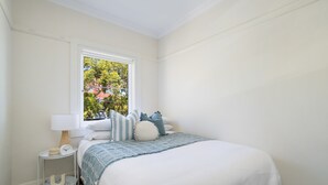 A bedroom with a bed, a window, a bedside table with a lamp, and a small shelf. - Little Manly 3 bedroom Oasis (Manly)