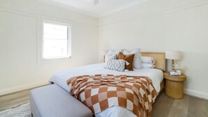 A bedroom with a bed, a bedside table, a lamp, and a window. - Little Manly 3 bedroom Oasis (Manly)