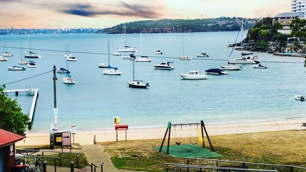 A marina with multiple boats, a beach with a swing set, and a distant city skyline. - Little Manly 3 bedroom Oasis (Manly)
