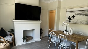 A living room with a fireplace, a flat-screen TV, a dining table with chairs, and a painting on the wall. - Little Manly 3 bedroom Oasis (Manly)
