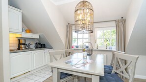 A kitchen with white cabinets, a coffee maker, and a kettle. There is a dining table with chairs and a chandelier above it. - Roseneck App. 6 - Cozy vacation apartment for up to 2 persons with (Sylt-Ost)