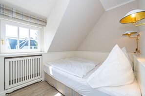 Quarto com teto inclinado, janela com vista para uma casa, radiador e luminária de cabeceira.