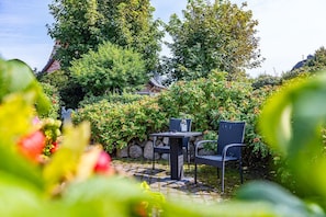 Um pátio com jardim, com mesa e cadeiras, rodeado por vegetação exuberante e arbustos floridos.