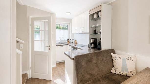 A compact kitchen with white cabinets, a coffee maker, and a dining area with a brown sofa and a cushion with text.