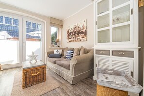 Ein gemütliches Wohnzimmer mit einem beigefarbenen Sofa, einem Holztisch und einem Korbwannenstuhl. Ein großes Fenster bietet Blick auf ein Gebäude.