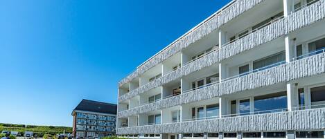 Un hotel de varios pisos con balcones, un camino de entrada pavimentado y un cielo azul despejado.