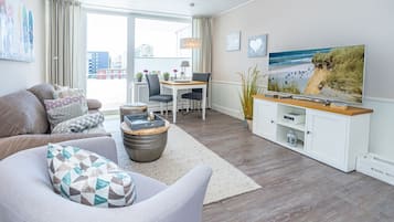 Ein modernes Wohnzimmer mit Sofa, Couchtisch und Schreibtisch mit Computer. Ein großes Fenster mit Vorhängen, ein Fernseher und Kunstwerke an der Wand.