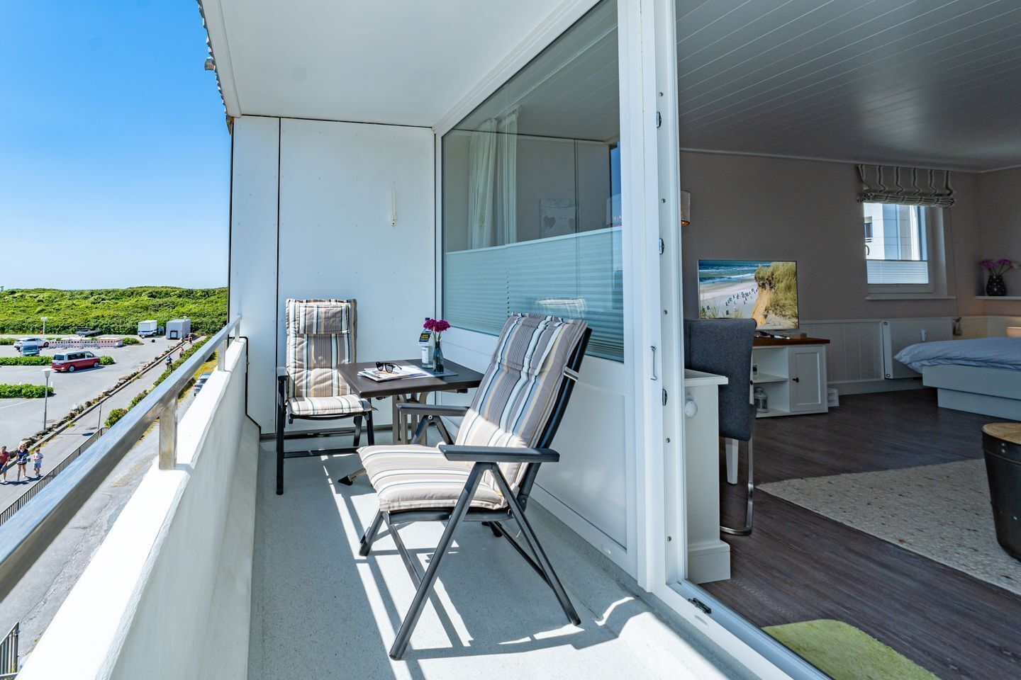 A balcony with two chairs, a small table, and a view of a parking area and greenery.
