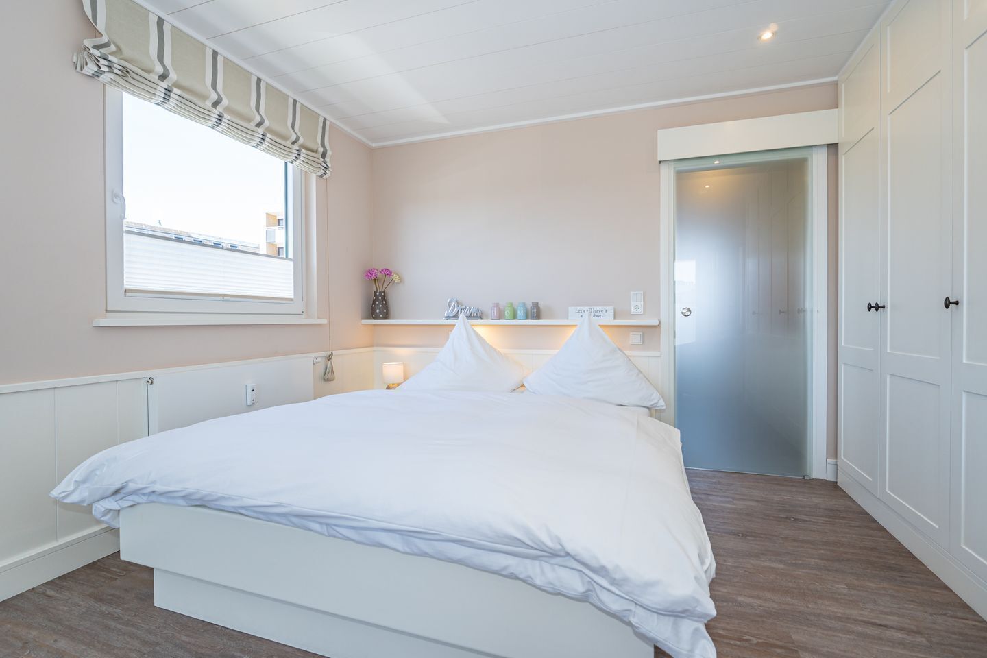 Ein Schlafzimmer mit Bett, Fenster mit Jalousien, Nachttisch mit Lampe, Regal mit Dekoration und einer Tür zu einem anderen Raum.