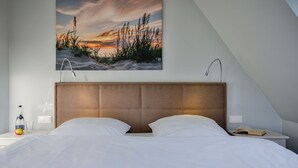Ein Schlafzimmer mit einem großen Bett, einem Kopfteil, einem Nachttisch mit einer Flasche und einem Glas sowie einem Gemälde eines Sonnenuntergangs über einem Strand.