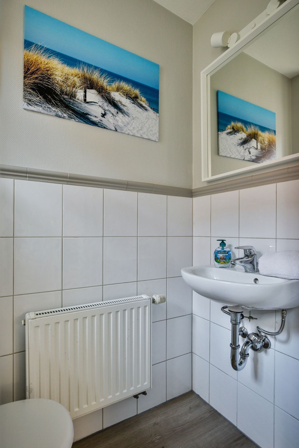 Une salle de bain avec un lavabo blanc, un miroir et un radiateur. Un tableau représentant une scène de plage est accroché au mur.