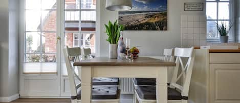 Un coin repas avec une table en bois et des chaises blanches, un luminaire suspendu, une plante en pot et un tableau encadré au mur.