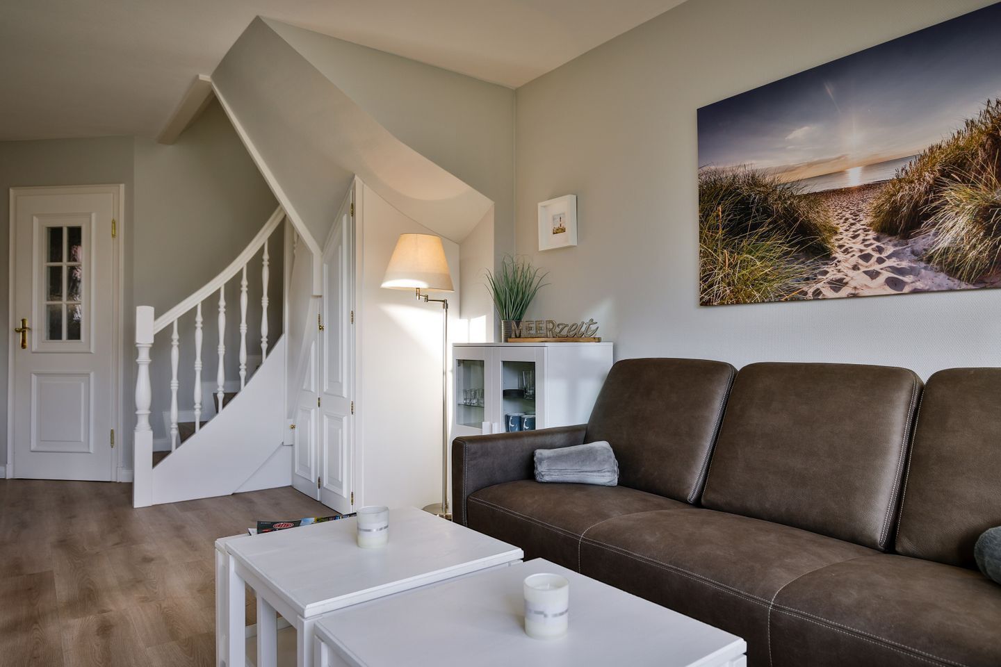 A living room with a brown leather sofa, a white coffee table, a lamp, and a painting of a beach scene.