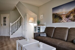 Un salon avec un canapé en cuir marron, une table basse blanche, une lampe et un tableau représentant une scène de plage.