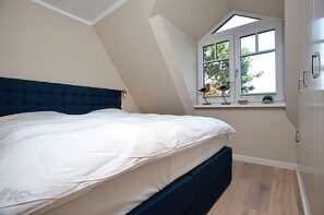 A bedroom with a large bed, a sloped ceiling, a window with a view of trees, and a small shelf with decorative items.
