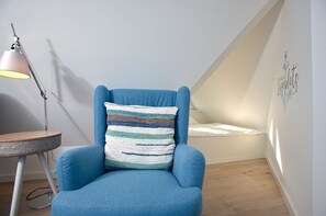 A blue armchair with a striped cushion, a small round table with a lamp, and a slanted ceiling with a bed. - Syltmuschel - Relaxen Sie in der Wunderschönen und Neuen 3-zimmerwohnung in Westerland (Sylt)