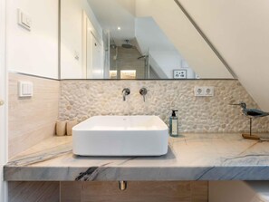 A bathroom with a large white sink, a marble countertop, a pebble-patterned wall, a bird sculpture, and a mirror reflecting a staircase. - Syltmuschel - Relaxen Sie in der Wunderschönen und Neuen 3-zimmerwohnung in Westerland (Sylt)