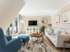 A modern living room with a white sofa, blue armchairs, a wooden coffee table, and a television on a white cabinet. - Syltmuschel - Relaxen Sie in der Wunderschönen und Neuen 3-zimmerwohnung in Westerland (Sylt)