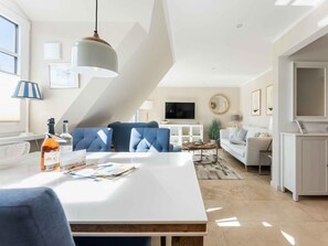 A modern living room with a white dining table, blue sofas, a TV, and a large window. - Syltmuschel - Relaxen Sie in der Wunderschönen und Neuen 3-zimmerwohnung in Westerland (Sylt)