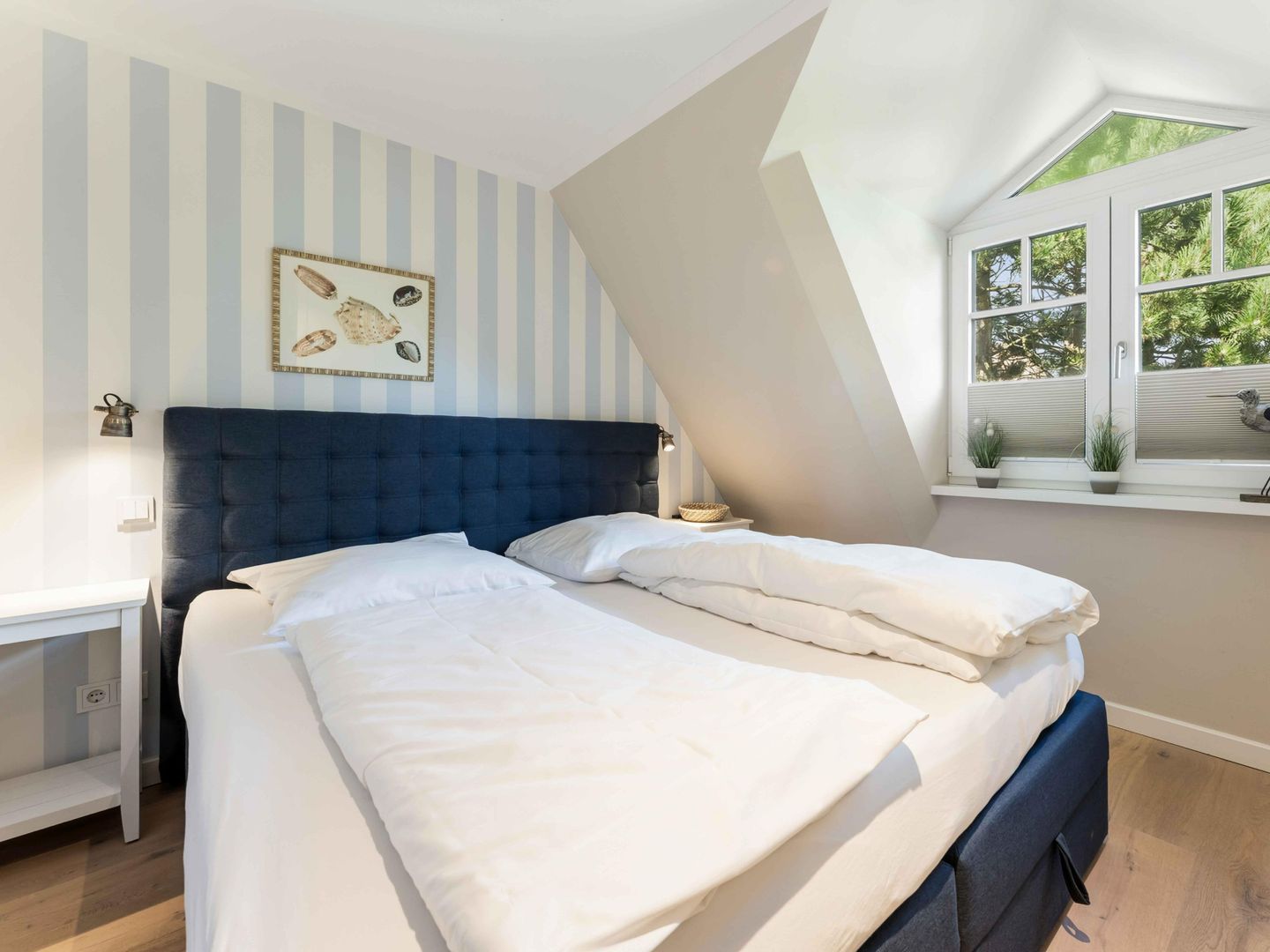 Een slaapkamer met een groot bed, een schuine kap, een raam met uitzicht op bomen, en een muur met gestreept behang en een lijst met een afbeelding.