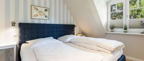 Een slaapkamer met een groot bed, een schuine kap, een raam met uitzicht op bomen, en een muur met gestreept behang en een lijst met een afbeelding.
