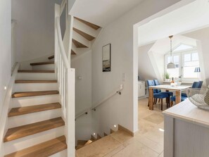 A modern interior with a wooden staircase, a dining area with blue chairs, and a kitchen in the background. - Syltmuschel - Relaxen Sie in der Wunderschönen und Neuen 3-zimmerwohnung in Westerland (Sylt)
