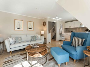 A modern living room with a sofa, armchair, and coffee table. A staircase leads to a kitchen area. - Syltmuschel - Relaxen Sie in der Wunderschönen und Neuen 3-zimmerwohnung in Westerland (Sylt)