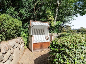 A small, enclosed outdoor seating area with a striped awning, surrounded by dense foliage and a stone wall. - Syltmuschel - Relaxen Sie in der Wunderschönen und Neuen 3-zimmerwohnung in Westerland (Sylt)
