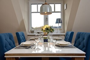 Mesa de jantar posta para quatro, com cadeiras estofadas em azul, um vaso de flores e uma garrafa de vinho.