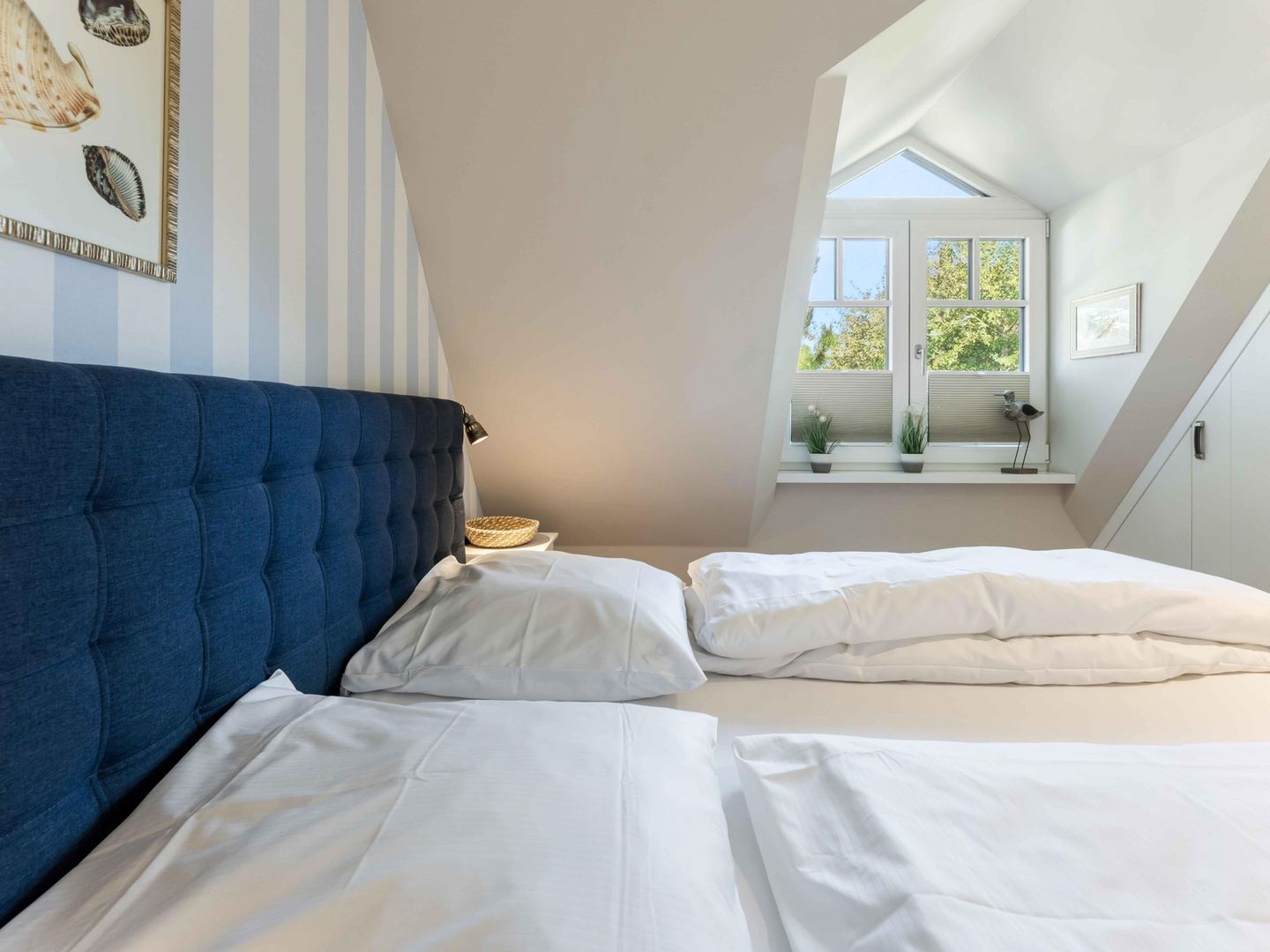 A bedroom with a blue headboard, two white beds, a small table with a lamp, and a window with a view of trees.
