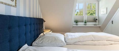 Une chambre à coucher avec une tête de lit bleue, deux lits blancs, une petite table avec une lampe et une fenêtre donnant sur des arbres.