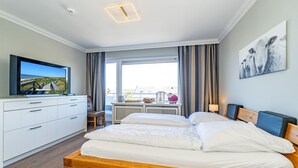A hotel room with a wooden bed, a TV on a white cabinet, a window with curtains, and a painting on the wall. - Adlerhorst - Modern vacation apartment with large roof terrace and private sauna in (Sylt)