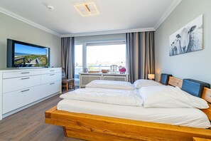 Habitación de hotel con una cama de madera, un televisor sobre un mueble blanco, una ventana con cortinas y un cuadro en la pared.
