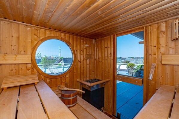 A wooden sauna room with a circular window, wooden benches, a sauna stove, and a view of buildings and a tower outside.