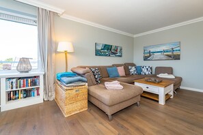 A cozy living room with a sectional sofa, a coffee table, a bookshelf, and a large window with curtains.