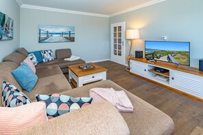 A living room with a sofa, coffee table, and TV stand. There is a painting on the wall and a door leading outside.