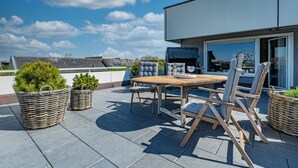 A rooftop patio with a wooden table and chairs, wicker planters with greenery, and a view of the cityscape. - Adlerhorst - Modern vacation apartment with large roof terrace and private sauna in (Sylt)