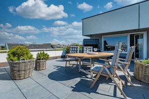 A rooftop patio with a wooden table and chairs, wicker planters with greenery, and a view of the cityscape.