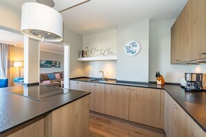 Una cocina moderna con armarios de madera, encimera negra y estufa integrada. En la encimera, un lavamanos con grifo, una cafetera y un bloque para cuchillos. Una lámpara está fijada a la pared sobre el lavamanos, y hay un plato decorativo con texto en la pared.