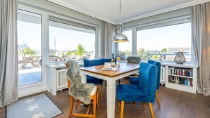 A dining area with a wooden table, blue chairs, and a view of the outdoors through large windows. - Adlerhorst - Modern vacation apartment with large roof terrace and private sauna in (Sylt)