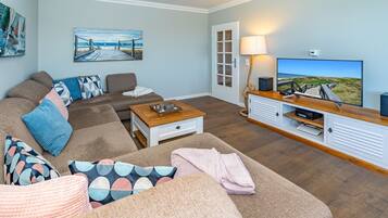 Ein Wohnzimmer mit Sofa, Couchtisch und TV-Standfuß. Ein Gemälde hängt an der Wand und eine Tür führt nach draußen.