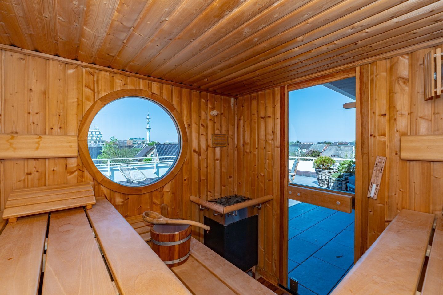 A wooden sauna room with a circular window, wooden benches, a sauna stove, and a view of buildings and a tower outside.