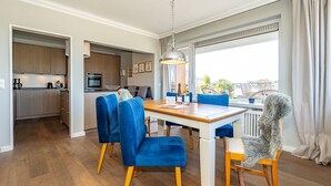 A modern kitchen with wooden cabinets, a built-in oven, and a dining area with a wooden table and blue upholstered chairs. - Adlerhorst - Modern vacation apartment with large roof terrace and private sauna in (Sylt)
