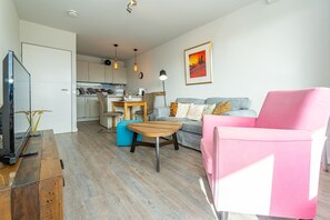 Ein modernes Wohnzimmer mit einem rosafarbenen Sessel, einem grauen Sofa und einem Holztisch. Im Hintergrund ist eine Küchenzeile mit Esstisch und Stühlen zu sehen.